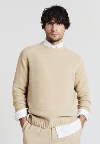 Beige Strickpullover mit rundem Halsausschnitt, geripptem Saum und Bündchen. Über einem weißen Hemd mit Kragen getragen, das eine strukturierte Oberfläche aufweist.