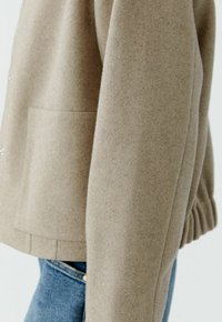 Hellbraune Wolljacke mit strukturiertem Finish, ausgestattet mit seitlicher Tasche und gerafften Bündchen. Neben blauen Denim-Jeans präsentiert.