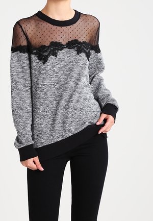 Grå langærmet sweater med gennemsigtig sort polkaprikket skulderstykke og blomsterblondekant, kombineret med sorte bukser på en stående person.