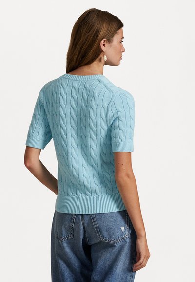 Frau in einem hellblauen, kurzärmeligen Pullover mit Zopfmuster und blauen Jeans, die mit dem Gesicht abgewandt steht und den Kopf zur Seite dreht, vor einem weißen Hintergrund.