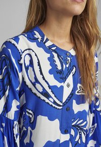 Blusa blu e bianca con motivo a fantasia, dotata di scollatura tonda, chiusura con bottoni e tessuto testurizzato con design botanico.