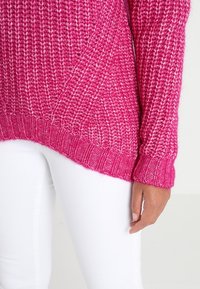 Maglione lavorato a maglia di un vivace colore rosa con un motivo testurizzato, con polsini e orlo a coste, abbinato a pantaloni bianchi.