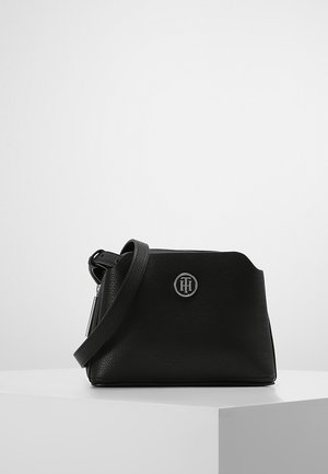 Sort læder crossbody taske med justerbar rem og sølvfarvet cirkulært logoemblem foran, vist på en hvid podium.
