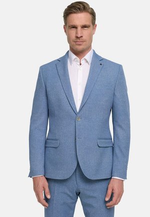 Blonder Mann in einem hellblauen Anzug mit weißem Hemd, steht frontal vor einem einfarbigen weißen Hintergrund.