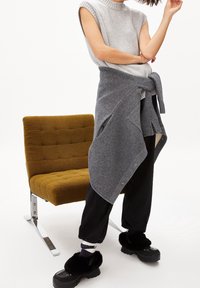 Szary bezrękawnik dzianinowy nałożony na szary szal zawiązywany; czarne spodnie zestawione z czarnymi, puszystymi butami wsuwanymi; obok znajduje się musztardowe pikowane krzesło.