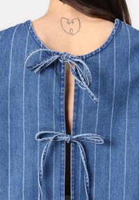 Indumento in denim con strisce verticali blu, caratterizzato da un collo rotondo e fiocchi sul retro, con un piccolo taglio sotto.