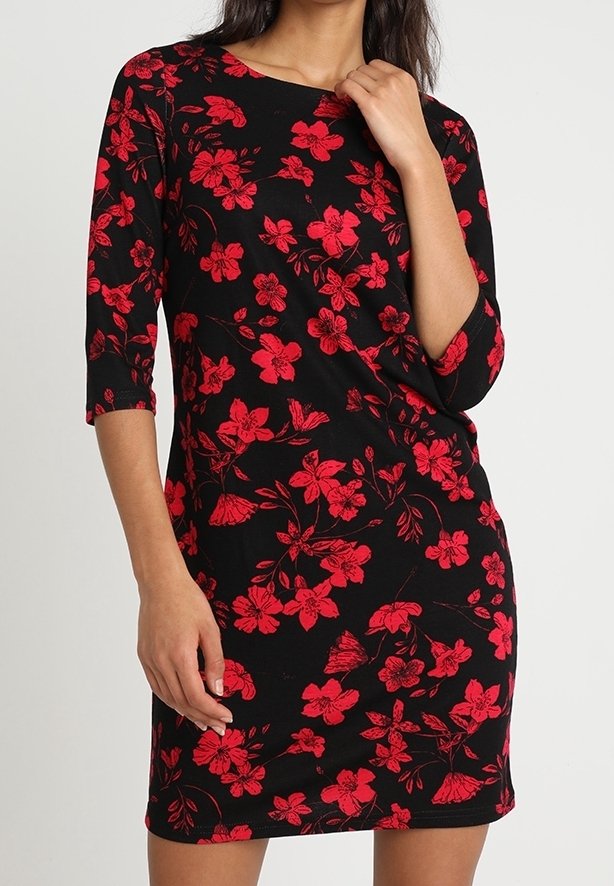Femme portant une robe noire mi-longue à imprimé floral rouge et manches trois-quarts, debout avec une main près du cou et l'autre le long du corps.
