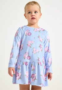 Młode dziecko o blond włosach, ubrane w długą, jasnoniebieską sukienkę z różowymi i fioletowymi kwiatowymi wzorami, stojące na jednolitym tle.
