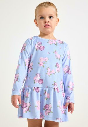 Malé dítě se světlými vlasy, oblečené v dlouhých rukávech světlemodrých šatů s růžovými a fialovými květinovými vzory, stojící před jednobarevným pozadím.