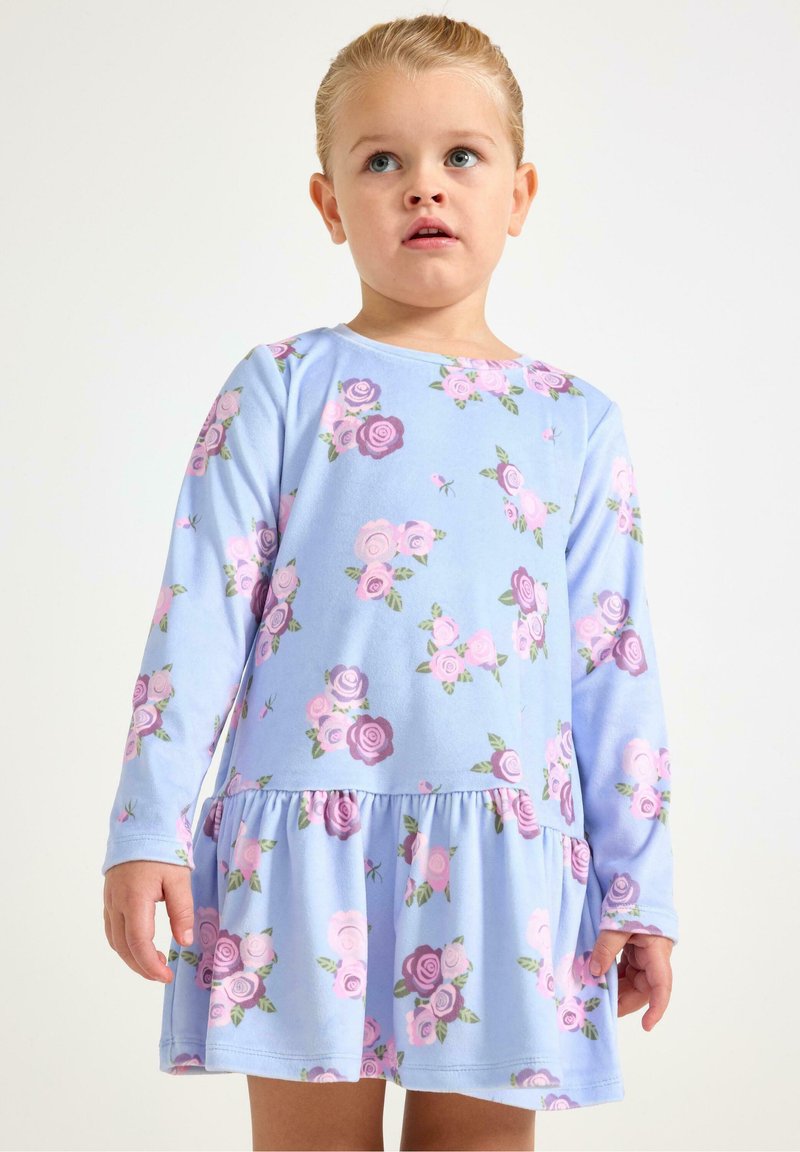 Młode dziecko o blond włosach, ubrane w długą, jasnoniebieską sukienkę z różowymi i fioletowymi kwiatowymi wzorami, stojące na jednolitym tle.