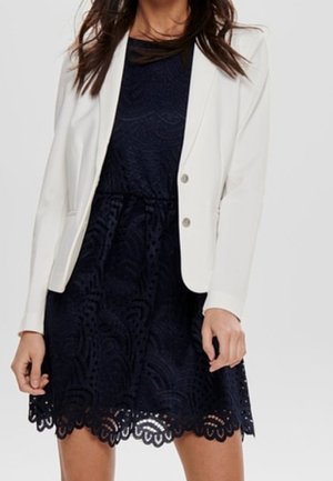 Vrouw die een marineblauwe kanten jurk draagt met een geschulpte zoom en een witte blazer met twee knopen eroverheen, staand tegen een effen achtergrond.