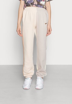 Femme portant un jogging Nike beige et crème avec des poignets élastiques et des baskets blanches, debout devant un fond gris uni.