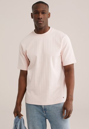 Man draagt een lichtroze geribd T-shirt met korte mouwen en lichtblauwe spijkerbroek, houdt een blauwe spijkerjack in één hand vast tegen een effen achtergrond.