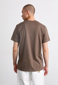 Brun bomulds t-shirt med korte ærmer, rund halsudskæring og lige kant. Synlig label på den nederste højre side. Model vendt om.