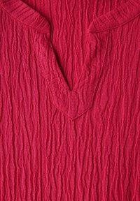 Blouse en tissu rouge froissé présentant un décolleté en V profond et une surface texturée, mettant en valeur des lignes verticales et un matériau doux et léger.