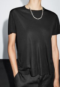 Sort top med korte ærmer i glat stof, med en afslappet pasform og rund halsudskæring, kombineret med en sølvfarvet perlehalskæde.