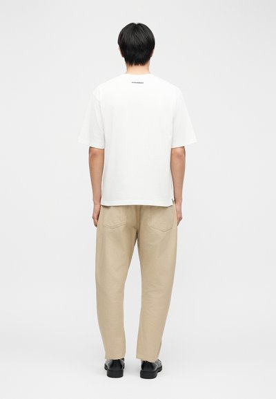 Άνδρας που φοράει λευκό κοντομάνικο μπλουζάκι και μπεζ παντελόνι, στέκεται με την πλάτη προς τα εμπρός σε απλό γκρι ανοιχτό φόντο.