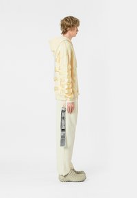 Sudadera con capucha en amarillo pálido con acentos de cinta texturizada en las mangas, combinada con pantalones crema que tienen una correa estampada. Usada con calzado voluminoso.
