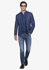 Marine ternet blazer, lagd over en blå sweater, parret med lyseblå jeans og sorte loafers. Accessories inkluderer solbriller.