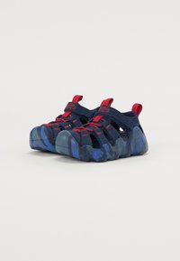 Zapatos acuáticos en azul y marino con suela texturizada, parte superior de malla, detalles en rojo y correas ajustables para un ajuste seguro. Patrón de camuflaje en la suela.