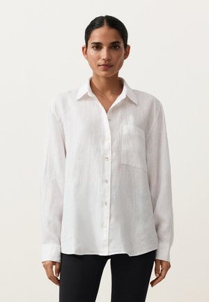 Frau mit zurückgebundenem dunklem Haar, die ein locker sitzendes weißes Hemd mit Brusttasche und schwarze Hosen trägt, vor einem einfarbigen Hintergrund.