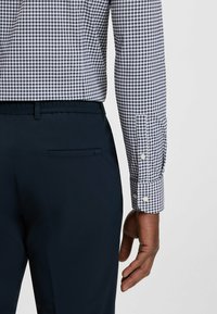 Sininen ja valkoinen ruudullinen paita, jossa on nappihihansuut, yhdistetty tummansinisiin housuihin, joissa on takatasku läpällä. Tekstuurikangas näkyvissä.