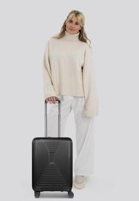 Donna bionda in dolcevita beige e pantaloni bianchi, in piedi con una mano sulla maniglia di una valigia rigida nera con rotelle.