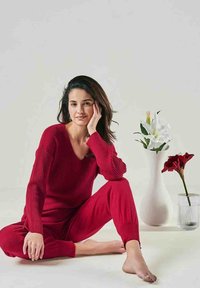 Roter gerippter Strickpullover mit V-Ausschnitt und dazu passenden Hosen. Barfußmodell sitzt neben weißen und roten Blumen in Vasen vor einem neutralen Hintergrund.