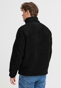 Black fleece jacket with a high collar, textured surface, and relaxed fit. Features long sleeves and a straight hem, paired with blue jeans.