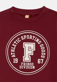 Felpa bordeaux con una grafica circolare bianca che dice "ATHLETIC SPORTING GOODS", con "FRIBOO DIVISION" e un logo "F".