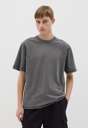 Camiseta de algodón gris con cuello redondo, corte relajado, mangas cortas y bordes deshilachados. Modelo de pie con una mano en el bolsillo, fondo neutro.