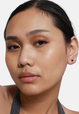 Boucles d'oreilles en studs noires carrées portées dans des oreilles percées. Surface en métal lisse, design minimaliste, contrastant avec le teint de la peau et la coiffure.