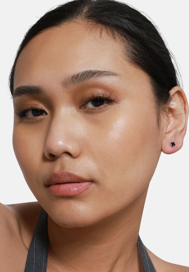 Orecchini a bottone neri quadrati indossati in orecchie forate. Superficie metallica liscia, design minimalista, in contrasto con il tono della pelle e acconciatura.