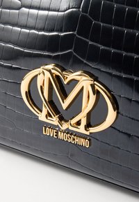 Fekete krokodilbőr mintázatú táska fényes felülettel, arany "Love Moschino" logóval és szív alakú fém díszítéssel.