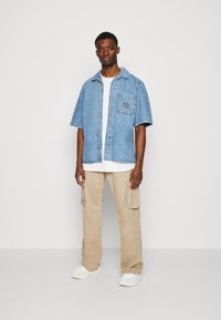 Gai zila džinsu krekls ar īsām piedurknēm, ar krūšu kabatu un logo, uzvilkts virs baltas T-krekla. Beigā krāsas bikses, baltas kedas.