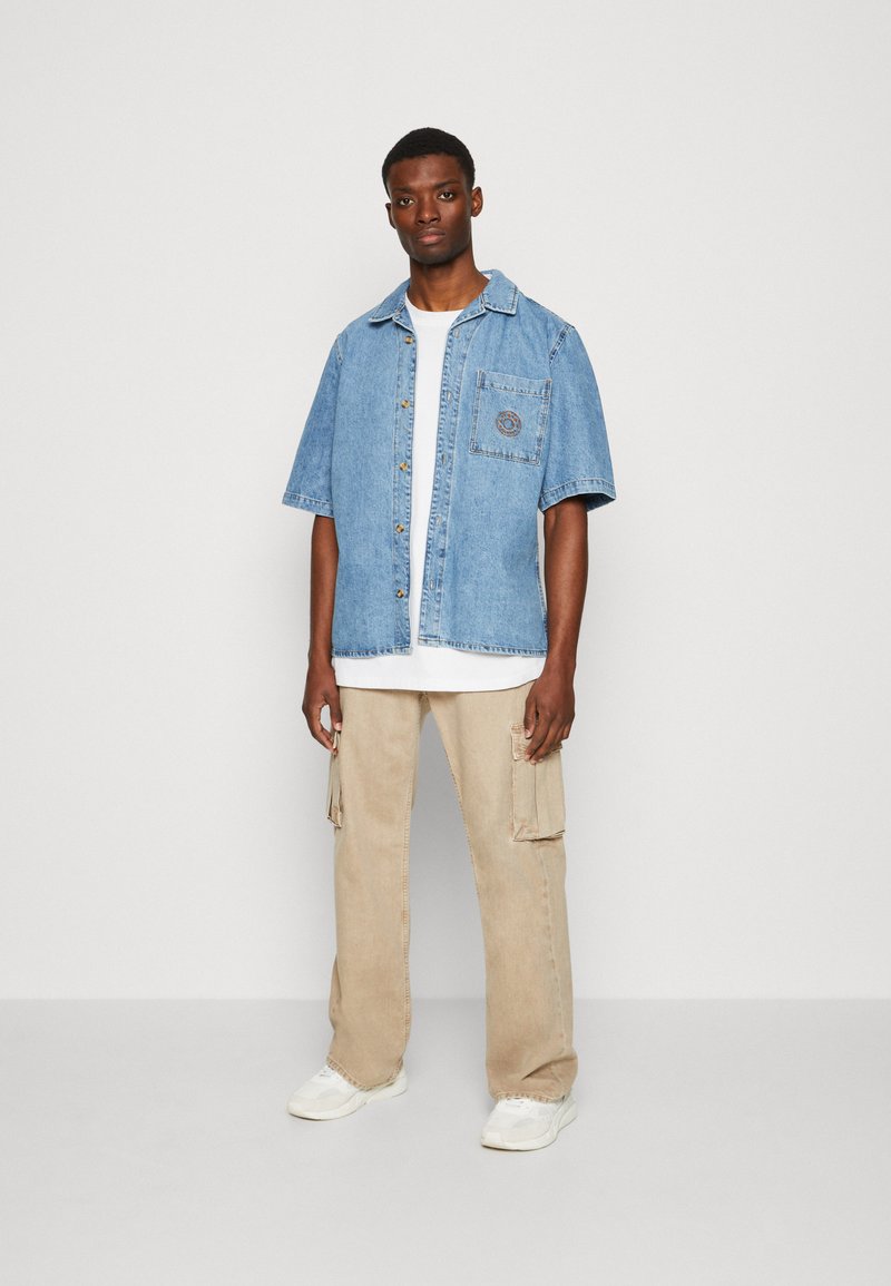 Gai zila džinsu krekls ar īsām piedurknēm, ar krūšu kabatu un logo, uzvilkts virs baltas T-krekla. Beigā krāsas bikses, baltas kedas.