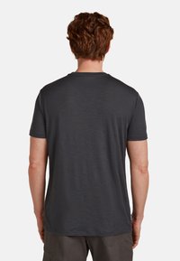 Hombre con cabello rojo rizado corto, vistiendo una camiseta de manga corta gris oscuro lisa y pantalones grises, de espaldas sobre un fondo blanco.