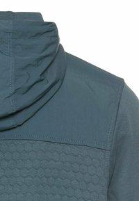 Veste à capuche avec un motif hexagonal texturé dans une couleur bleu tamisé, dotée d'une partie supérieure lisse et de détails de couture contrastants.
