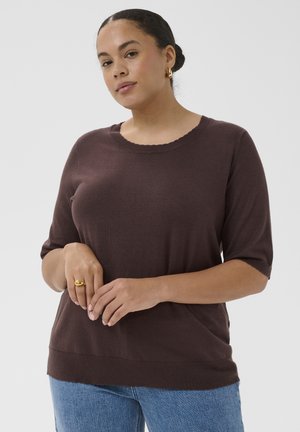 Femme aux cheveux foncés en chignon, portant un haut en maille marron à manches courtes, un jean bleu, des boucles d'oreilles anneaux dorés et une bague en or, debout devant un fond uni.