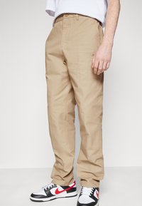 Beige chinos with a straight fit, featuring a stitched pocket and a small logo patch. Paired with black and white sneakers with red accents.