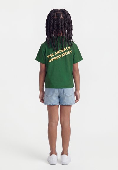 Παιδί με πλεξούδες, φοράει πράσινο T-shirt με την επιγραφή "THE ANIMALS OBSERVATORY", τζιν σορτς και λευκά αθλητικά παπούτσια, στέκεται με την πλάτη στραμμένη.