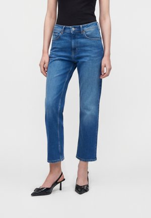 Kvinde iført blå jeans med lige ben, sort ærmeløs top og sorte slingback hæle med spids tå på en neutral baggrund.