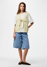 Femme debout, les mains sur les hanches, portant un gilet rayé jaune noué devant, un t-shirt blanc oversize, un short en jean large et des chaussures sombres sans lacets.