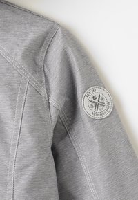 Manche de veste en tissu gris texturé avec un écusson blanc rond comportant des symboles et le texte « EST. 1981 » et « GLOBAL TRAVELER ».
