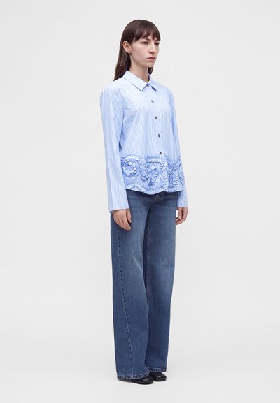 Femme portant une chemise à rayures bleues boutonnée avec des détails à volants floraux et un jean bleu à jambes larges, debout sur un fond blanc.