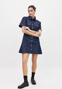 Tumedast sinisest denimist pluusikleit lühikeste varrukatega, nööpidega esiosal, kahe rinna-taskuga ja laiendatud seelikuosaga. Kandmisel mustade mokassiinide ja sokkidega.