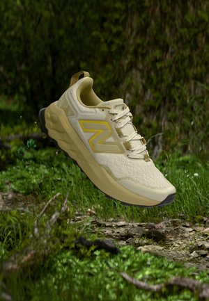 Chaussure de course New Balance beige avec des accents jaunes suspendue au-dessus d'un terrain herbeux et rocheux dans un cadre naturel en plein air.