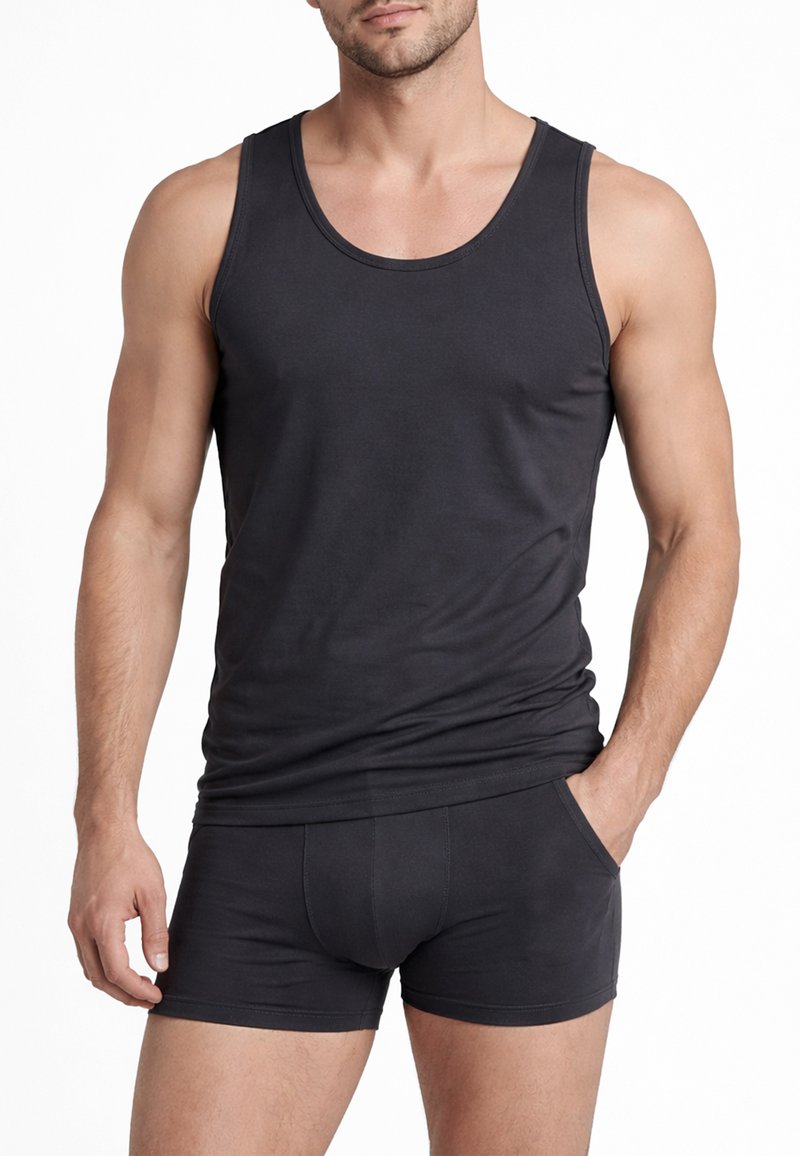 Uomo che indossa una canottiera nera aderente e boxer neri abbinati, in piedi con una mano in tasca contro uno sfondo bianco.