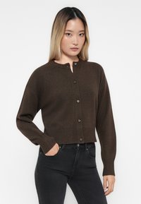 Bruine gebreide cardigan met een ronde halslijn, vijf knopen en ribbelmanchetten en -zoom. Draagt over strakke zwarte jeans.