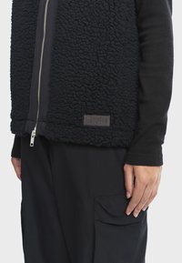 Čierna fleece vesta s textúrovaným, plyšovým povrchom, so strieborným zipsom. Nápis AIGLE na dolnom pravom rohu, nosí sa cez čierne nohavice.